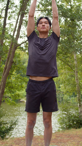 Video-Vertical-De-Un-Hombre-Vestido-Con-Ropa-Deportiva-Haciendo-Estiramientos-De-Yoga-Sobre-Una-Colchoneta-En-Un-Bosque-Junto-A-Un-Lago-O-Río-Disfrutando-De-La-Paz-Y-La-Belleza-De-La-Naturaleza-Filmado-En-Tiempo-Real-2